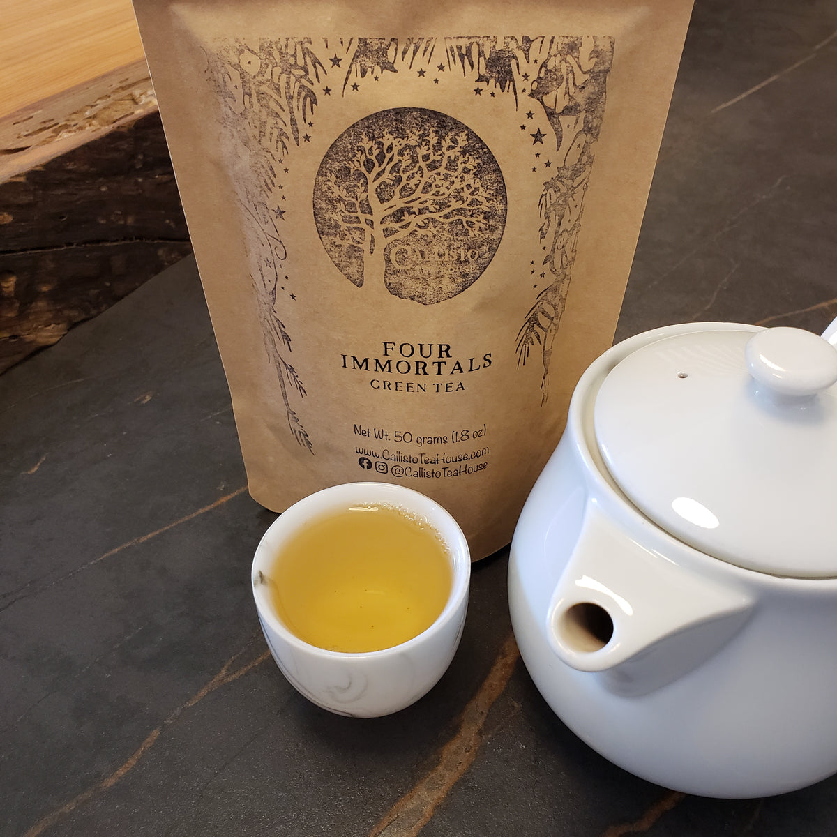 A kraft paper pouch of Four Immortals Green Tea stands on a dark stone surface next to a white teapot and a small white cup filled with brewed yellow-green tea.