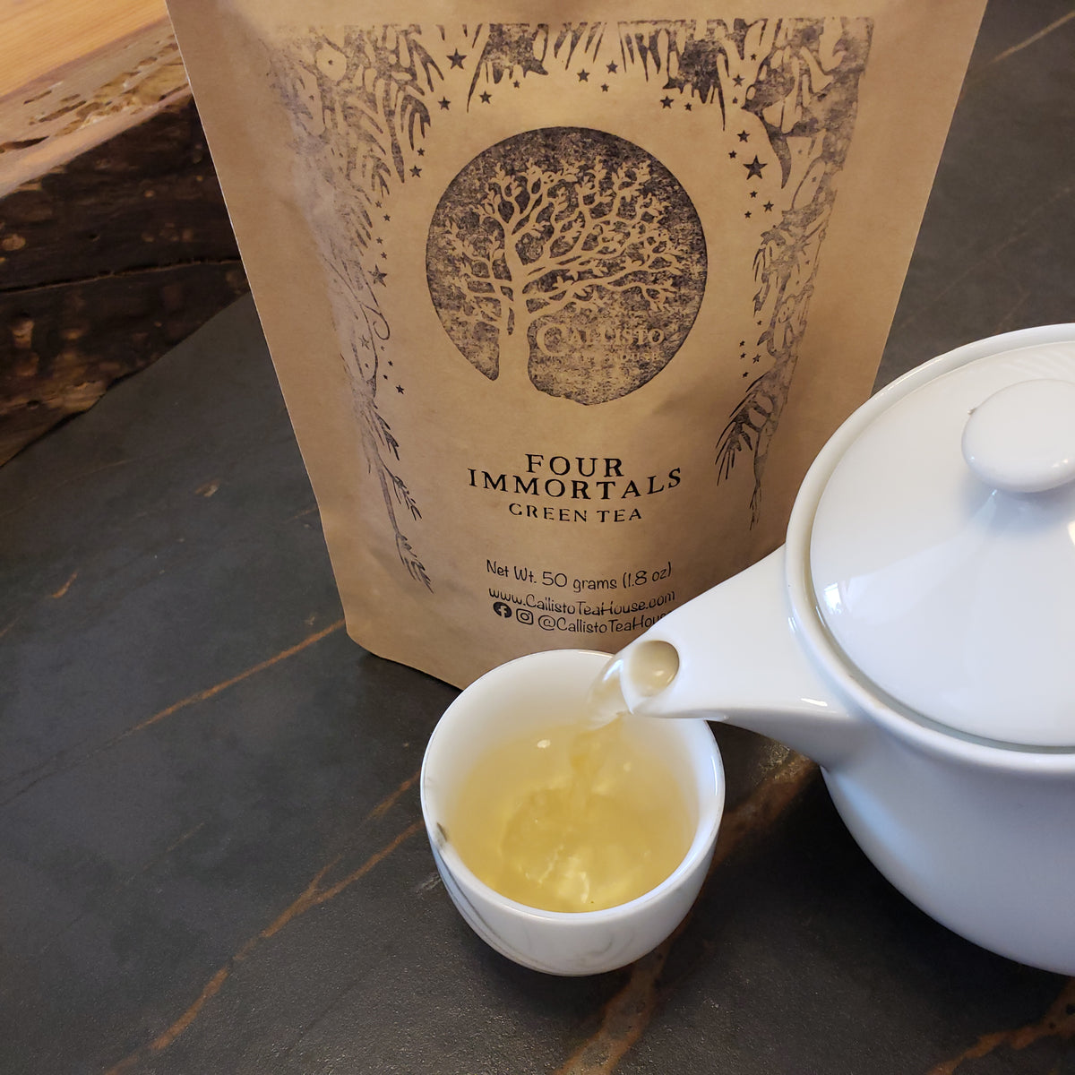 A kraft paper pouch of Four Immortals Green Tea stands on a dark tabletop beside a white teapot pouring pale gold tea into a small white cup.