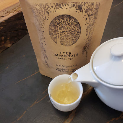A kraft paper pouch of Four Immortals Green Tea stands on a dark tabletop beside a white teapot pouring pale gold tea into a small white cup.
