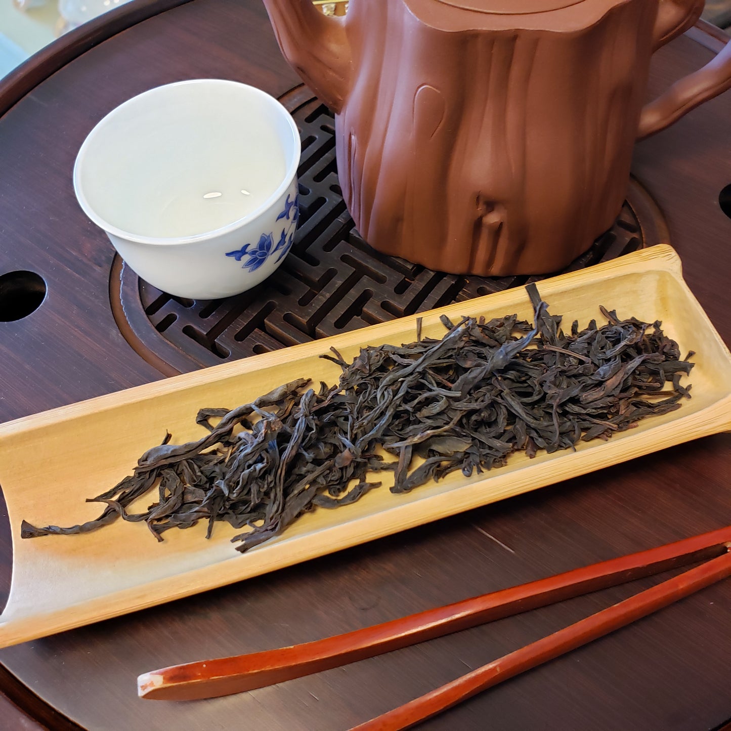 Tea leaves in a wooden tray with a teapot and cup on a wooden surface