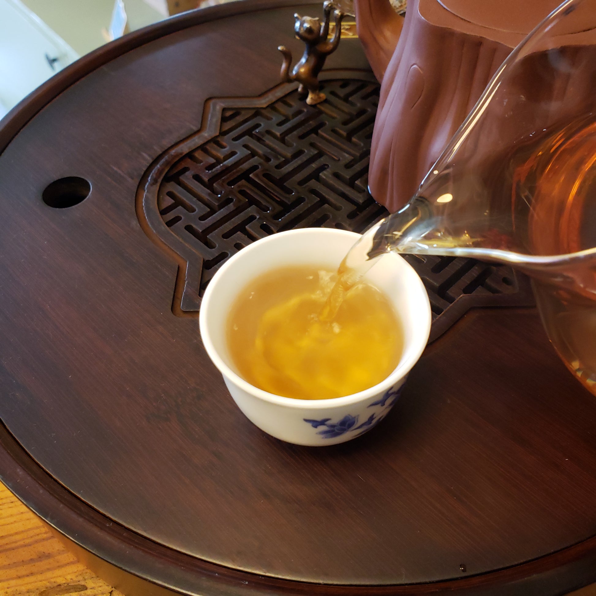Tea being poured from a glass teapot into a white ceramic cup on a wooden table.