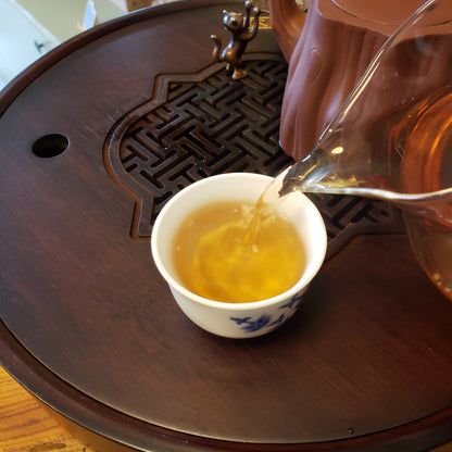 Tea being poured from a glass teapot into a white ceramic cup on a wooden table.