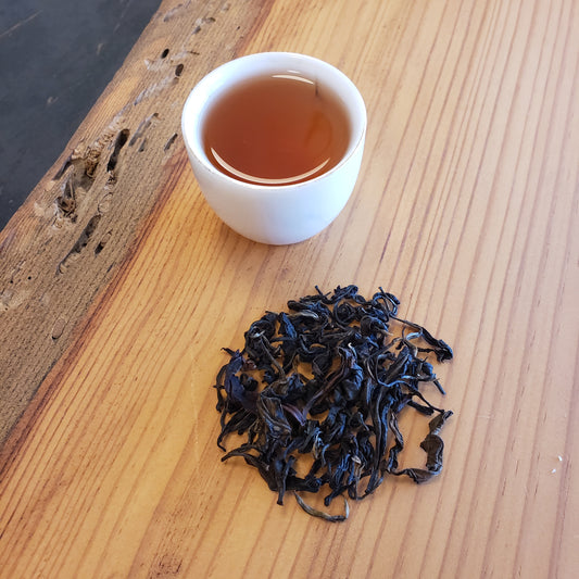 White cup of brewed tea next to a pile of black tea leaves on a wooden surface