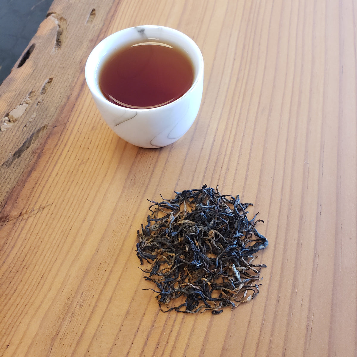A small white cup filled with amber-brown tea sits on a light wood table next to a small pile of loose-leaf tea buds.