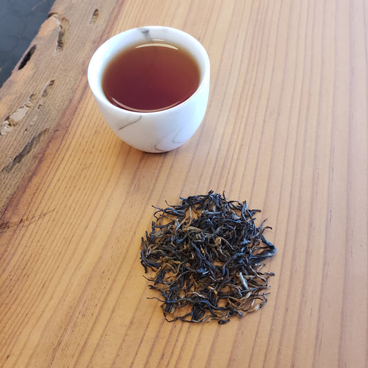A small white cup filled with amber-brown tea sits on a light wood table next to a small pile of loose-leaf tea buds.