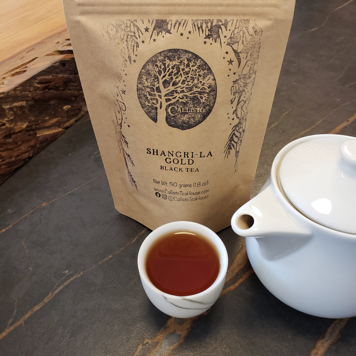 A kraft paper pouch of Callisto Tea House's Shangri-La Gold Black Tea stands on a dark stone surface next to a small white teacup filled with tea and a white teapot.