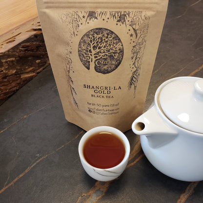 A kraft paper pouch of Callisto Tea House's Shangri-La Gold Black Tea stands on a dark stone surface next to a small white teacup filled with tea and a white teapot.