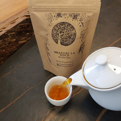 A stand-up kraft paper pouch of Shangri-La Gold Black Tea stands on a dark stone surface beside a white teapot pouring amber tea into a small white cup.