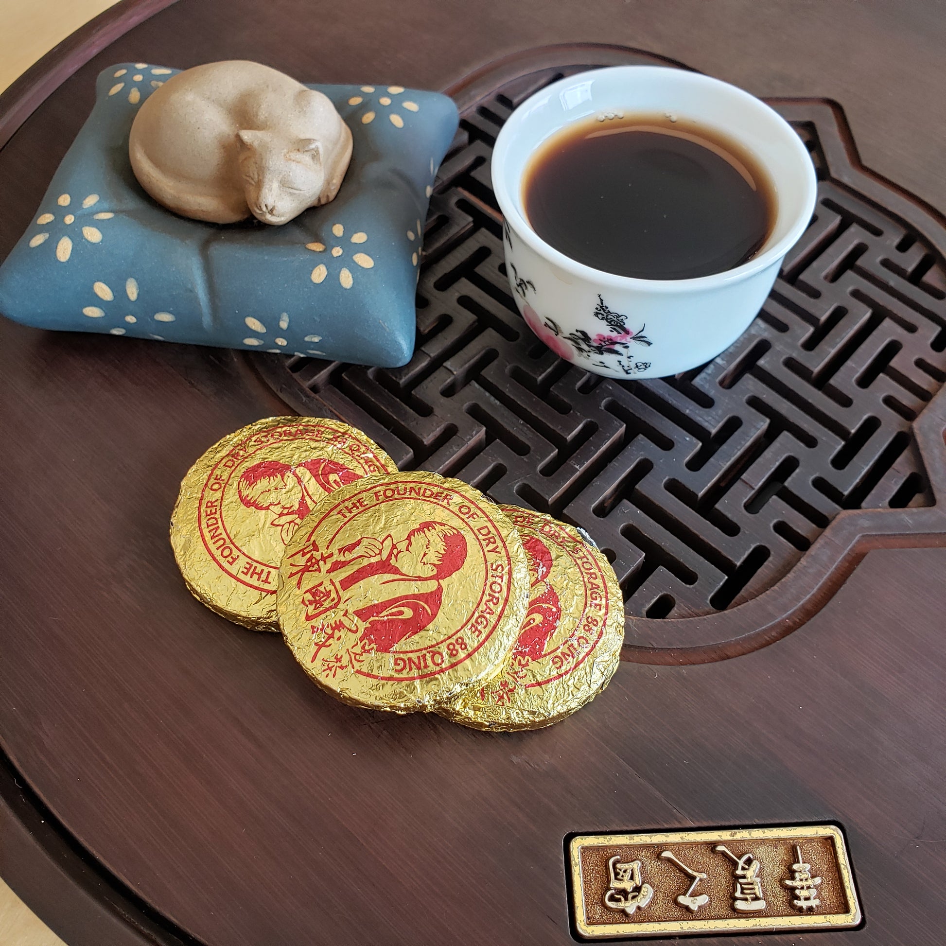 Tea cup with a decorative tray, ceramic cat tea pet, and gold tea leaves packaging on a wooden surface.