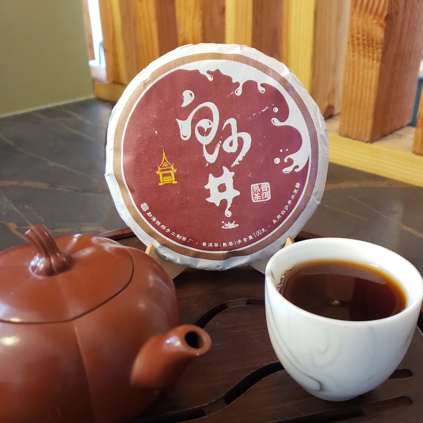 A round wrapped tea cake with a burgundy label sits on a wooden surface beside a brown teapot and a white cup of dark tea in a real indoor setting.