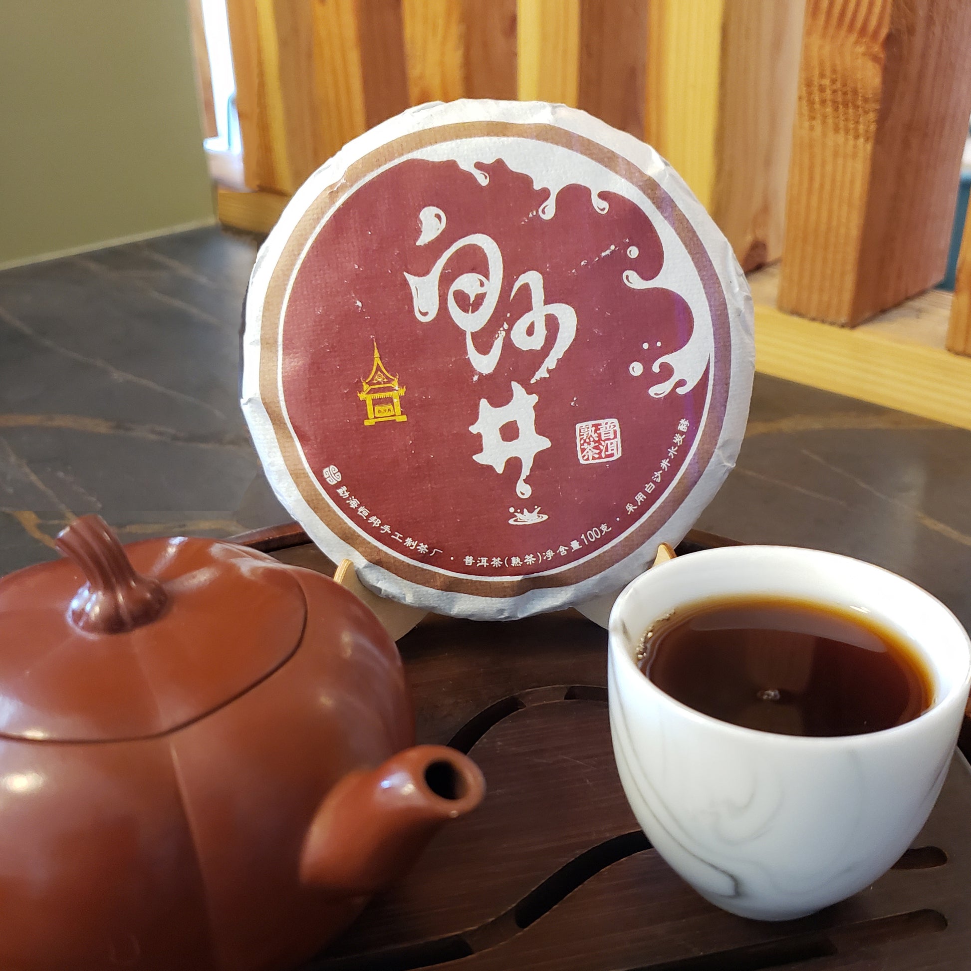 A round wrapped tea cake with a burgundy label sits on a wooden surface beside a brown teapot and a white cup of dark tea in a real indoor setting.