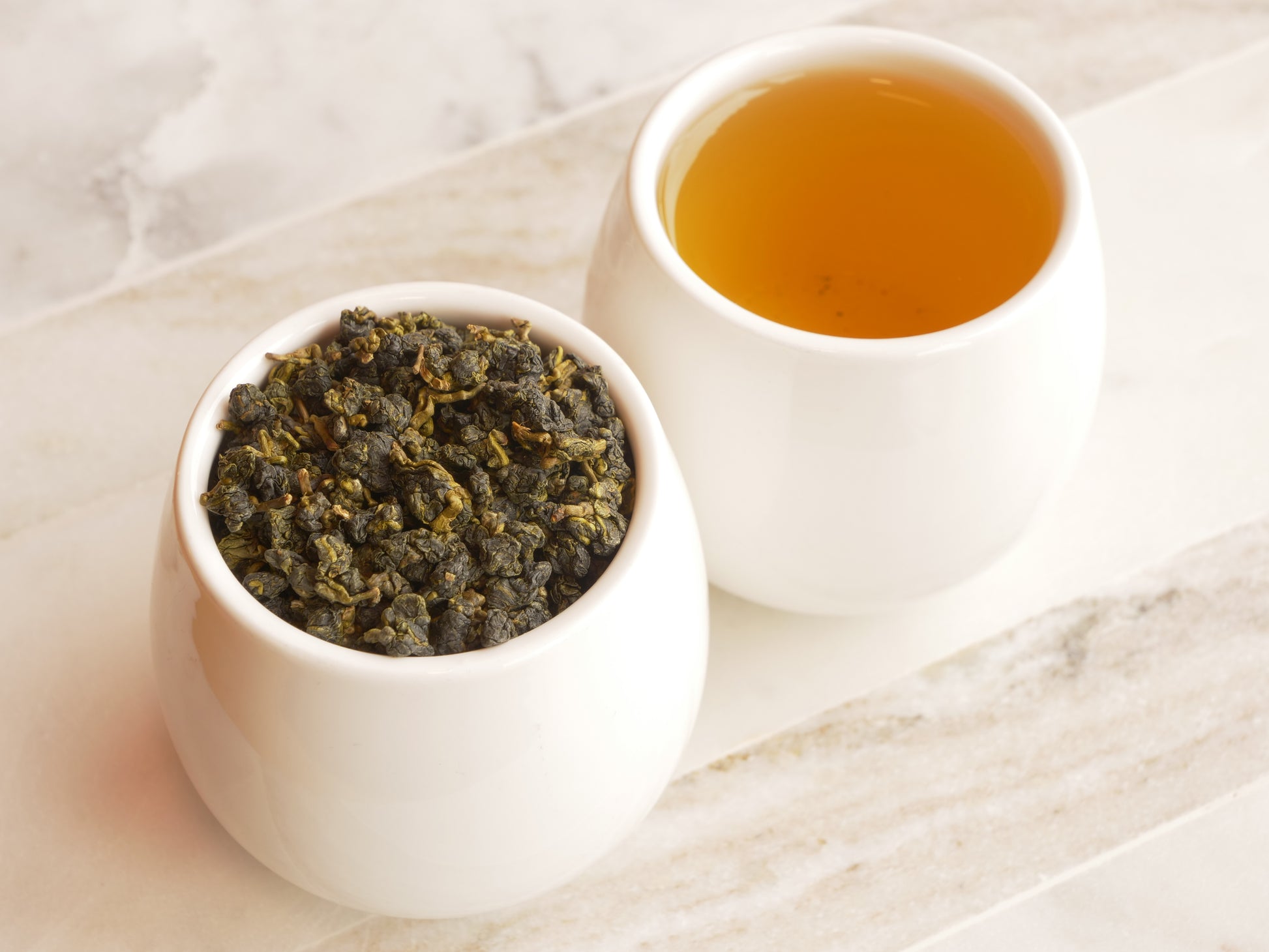 White bowl with tea leaves and a cup of brewed tea on a marble surface