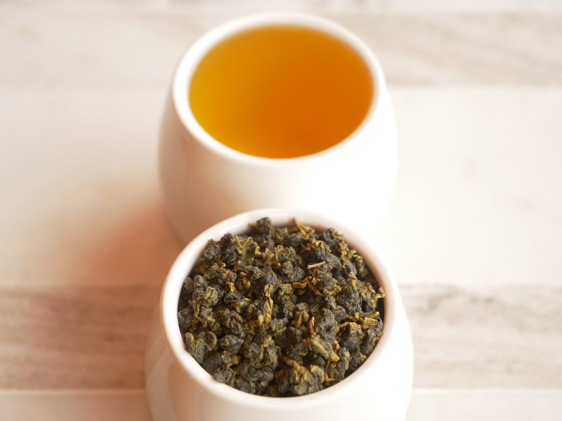 Two white bowls, one with dry tea leaves and the other with brewed tea, on a light wooden surface.