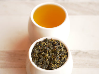 Two white bowls, one with dry tea leaves and the other with brewed tea, on a light wooden surface.