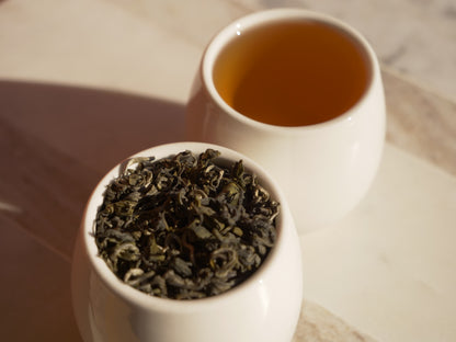 White cup with green tea leaves and a white cup with brewed green tea on a wooden surface
