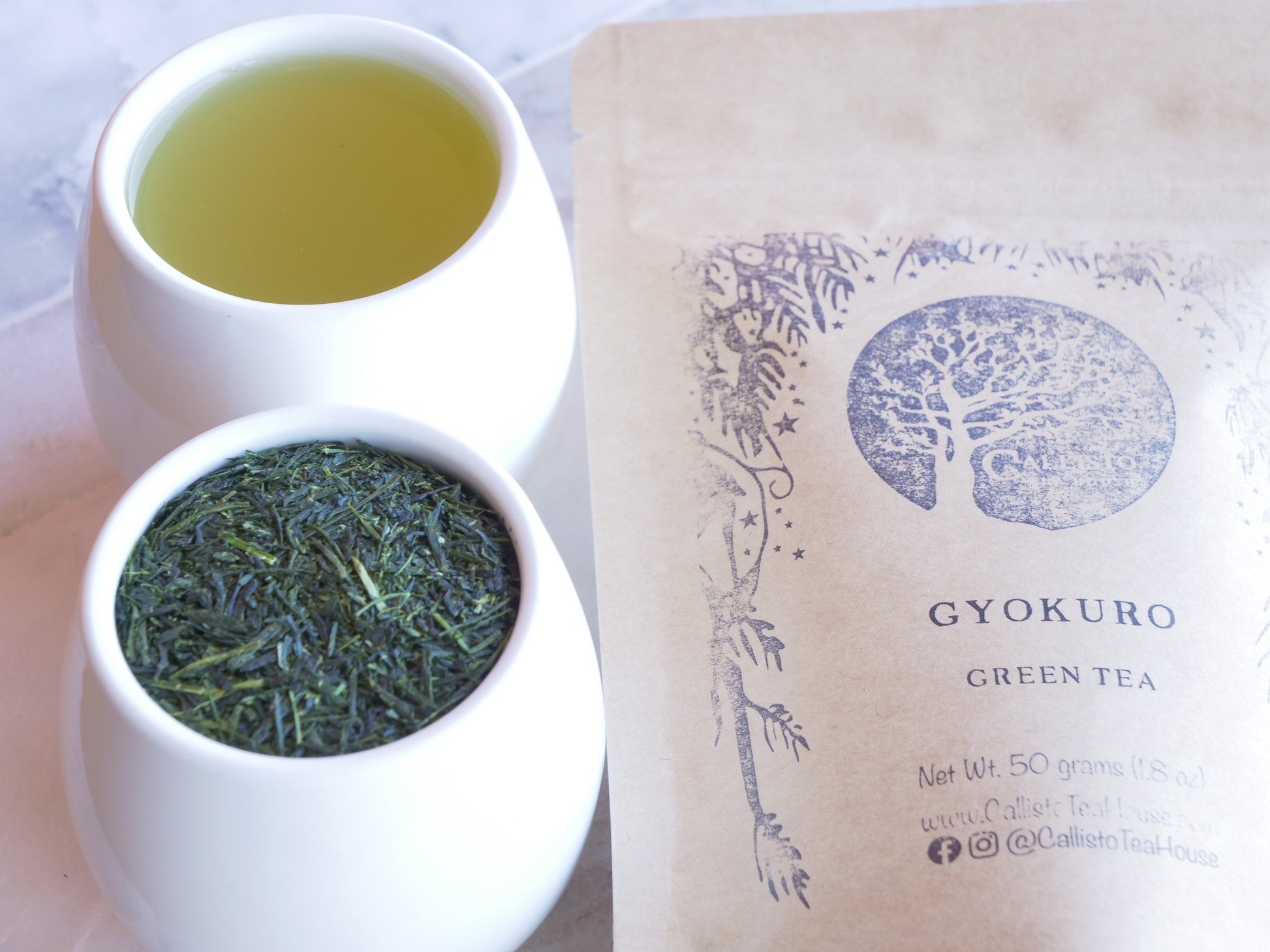 Two white containers with green tea leaves and a brown paper bag labeled 'Gyokuro Green Tea' on a light background.