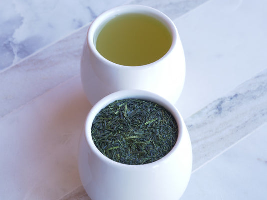 Two white ceramic containers with green tea leaves and brewed tea on a marble surface.