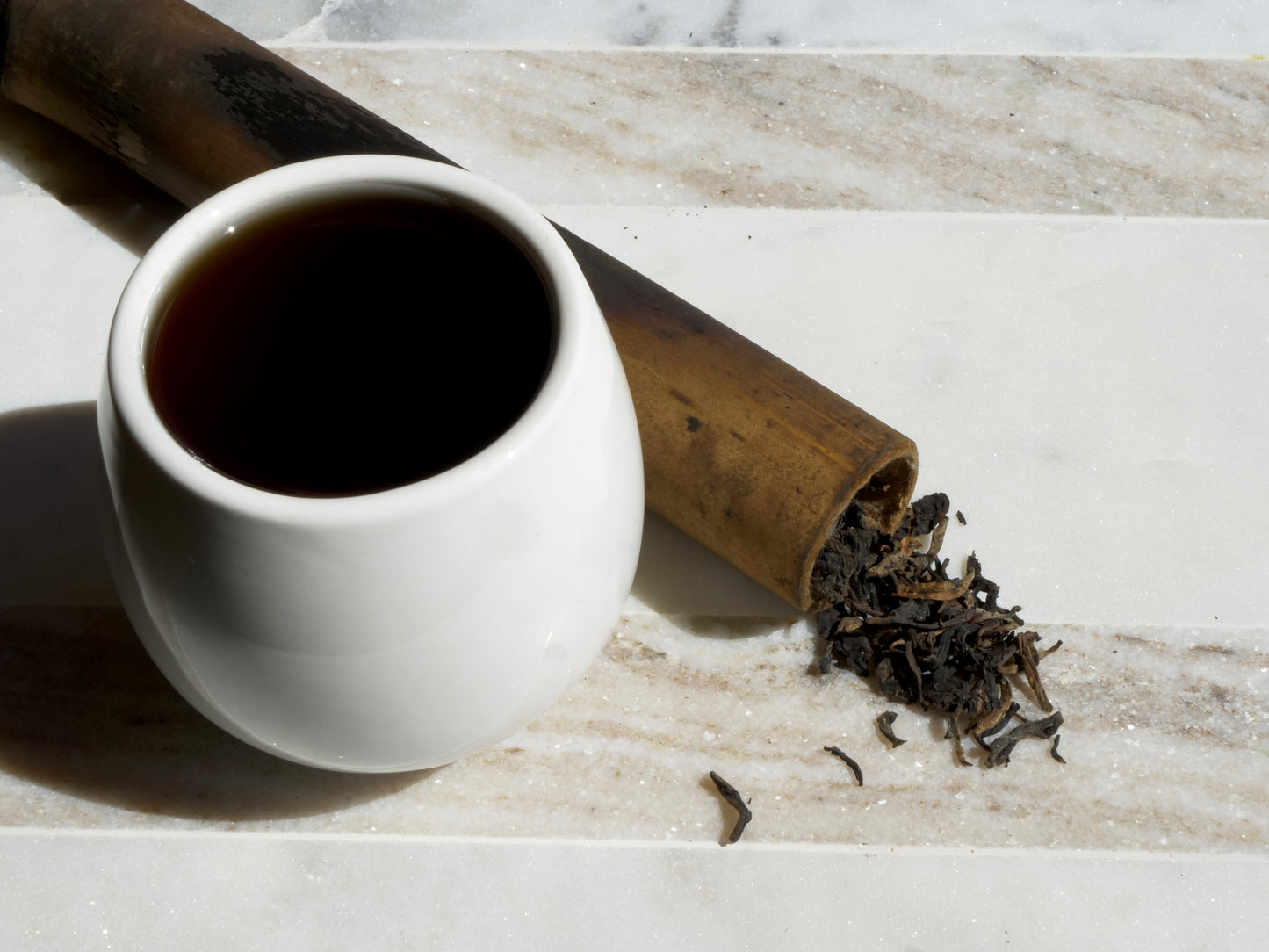 White ceramic cup filled with dark tea next to tea leaves and a bamboo tube on a marble surface