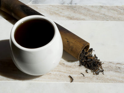 White ceramic cup filled with dark tea next to tea leaves and a bamboo tube on a marble surface